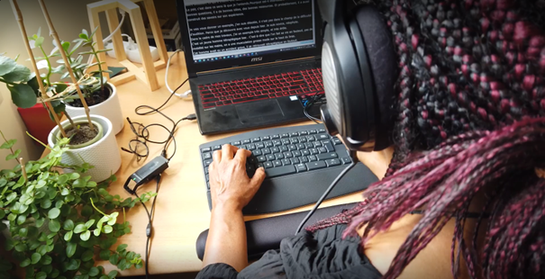 Une interprète de l'écrit de dos devant son bureau. Elle porte un casque audio. Un ordinateur est posé devant elle où s'affiche le sous-titrage.